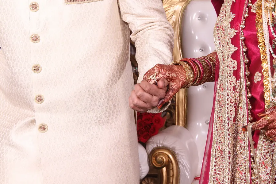 A groom in an ivory sherwani taking his bride's henna-decorated hand
