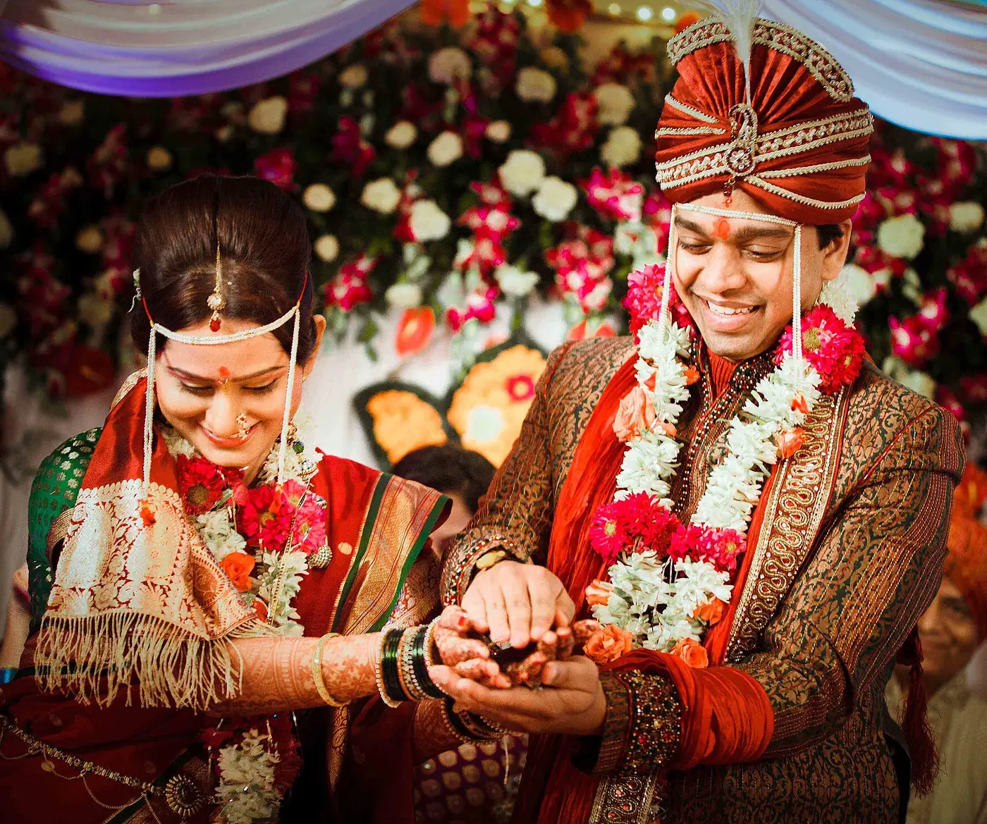 A smiling Indian bride and groom in traditional wedding attire and flower garlands, sharing a ritual during their wedding ceremony